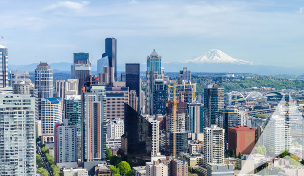 A panoramic view of downtown Seattle showcasing modern high-rise commercial and residential buildings with Mount Rainier visible in the distance under a clear blue sky. The image features Trinity Real Estate’s translucent white triangle icon overlay in the lower right corner, symbolizing the firm’s brand presence and commitment to urban real estate markets. Construction cranes are visible, highlighting ongoing development.